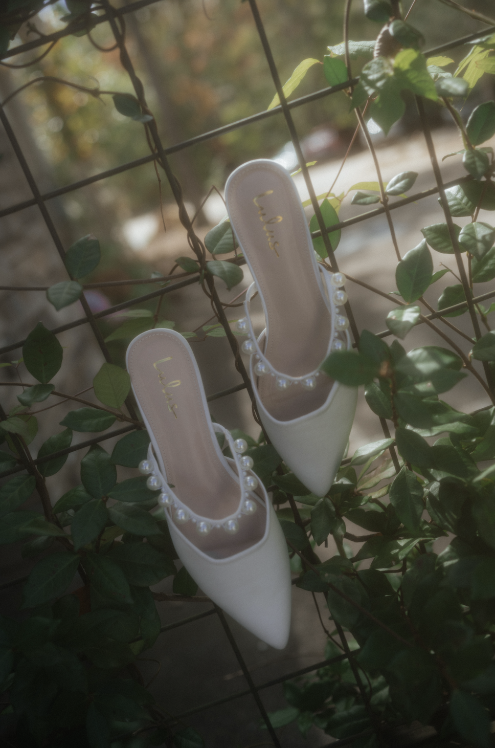 A pair of white high-heeled shoes with pearl embellishments, hanging on a grid against green leafy vines.