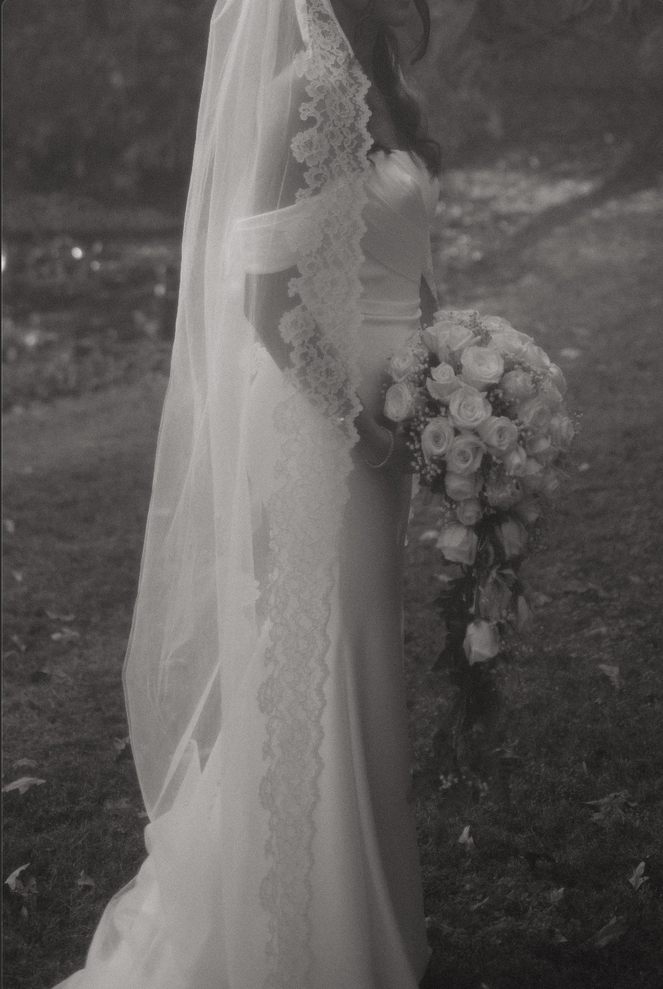 A bride holding a bouquet of roses, wearing a wedding dress and veil, standing outdoors on grass with fallen leaves.