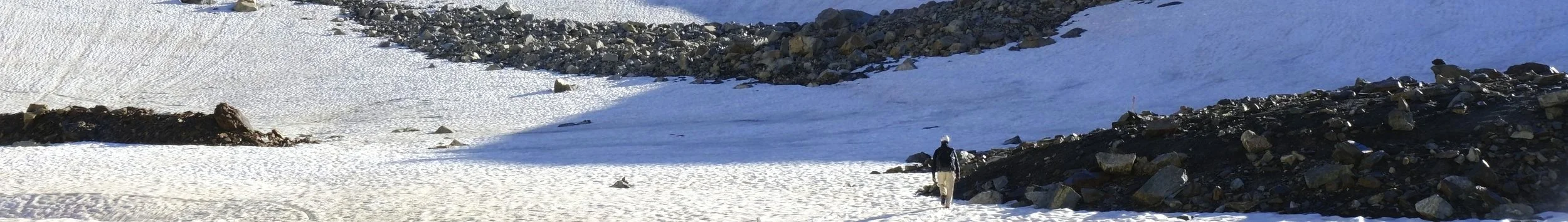 Ein Wanderer in Winterkleidung läuft durch eine verschneite Berglandschaft mit dunklen Felsen und schneebedeckten Hängen.