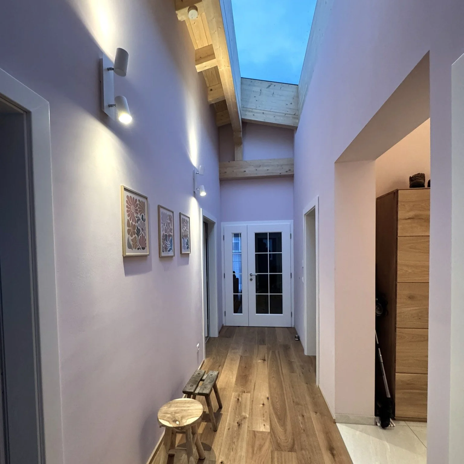 Ein Flur in einem modernen Haus mit Holzboden, Wandlampen, drei Bilder an der Wand, eine Glas-Tür am Ende und eine Holz-Schrank in einer Nische.