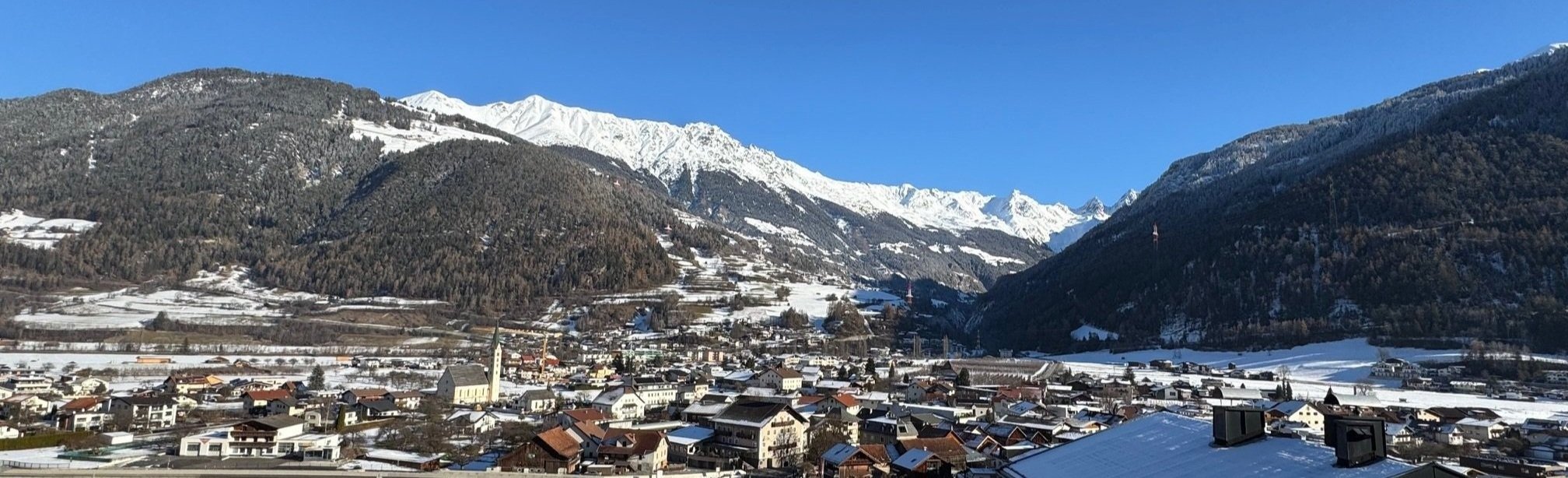 Bergdorf in einer winterlichen Gebirgsregion mit schneebedeckten Häusern vor hohen, schneebedeckten Bergen unter klarem blauen Himmel.