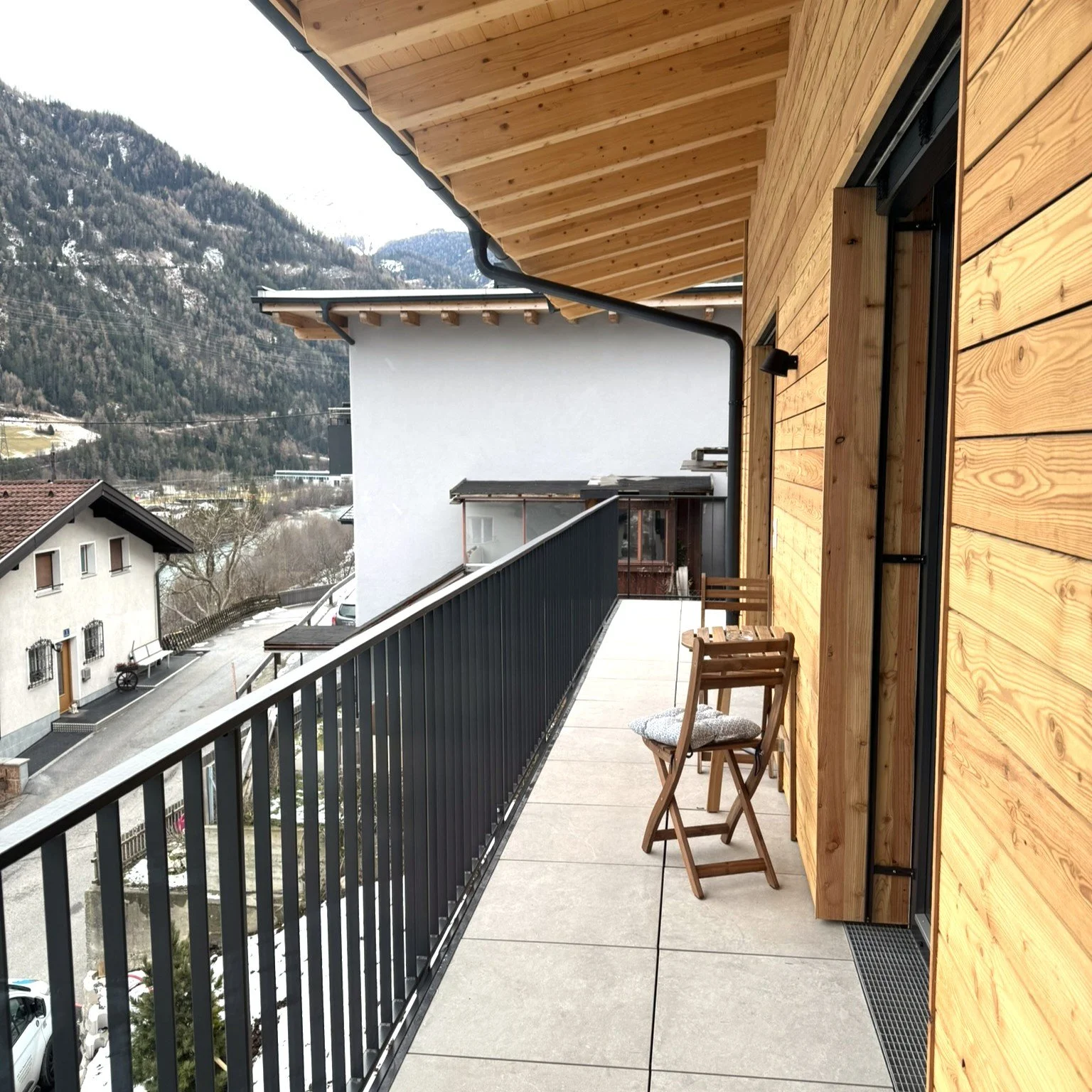 Blick auf einen Balkon mit Holzverkleidung, zwei Stühle, schwarzer Metallgeländer, in einer bergigen Gegend mit verschneiten Hügeln, Häuser, bewölkter Himmel.