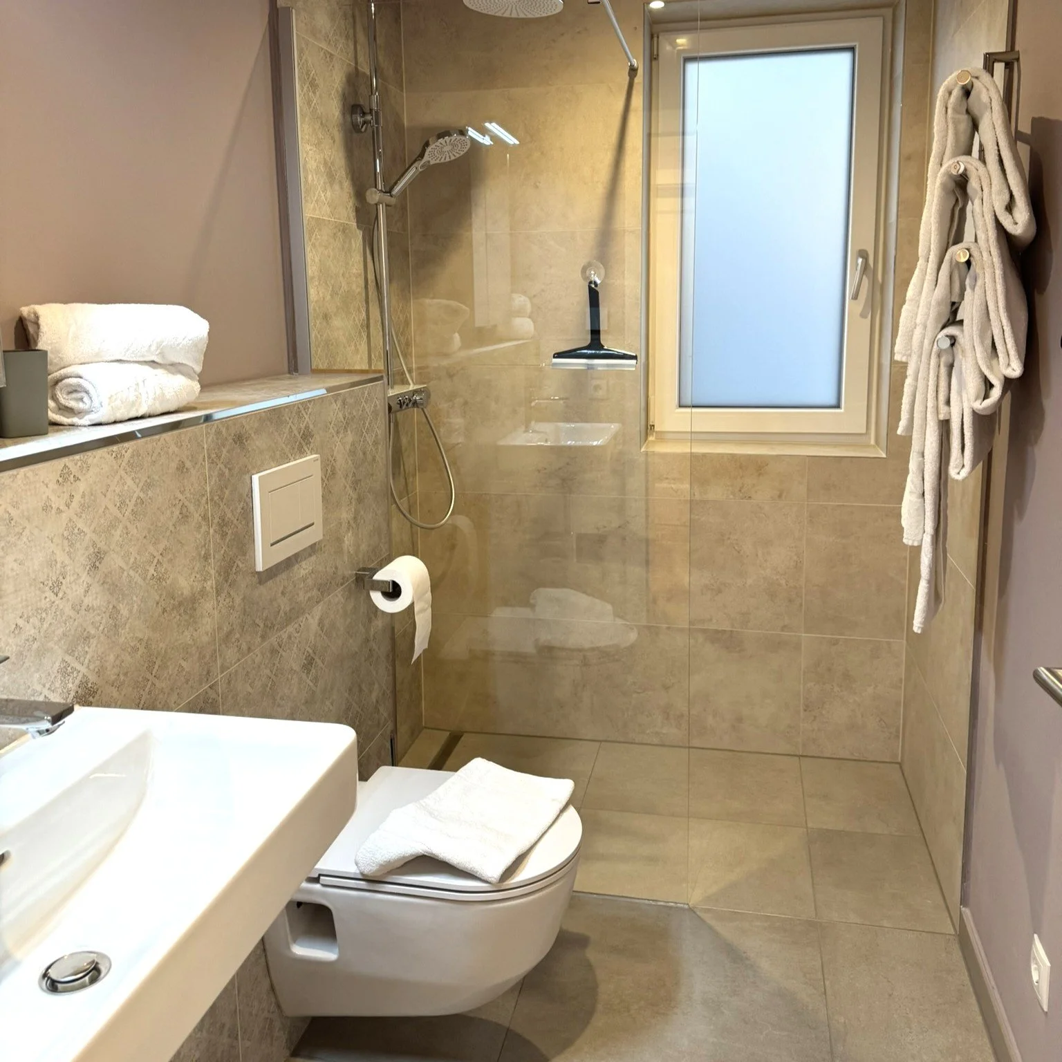 Ein modernes Badezimmer mit einer Toilette, Handtüchern, einem Waschbecken, einer Glas-Duschabtrennung, einem Fenster und Handtuchhaltern.