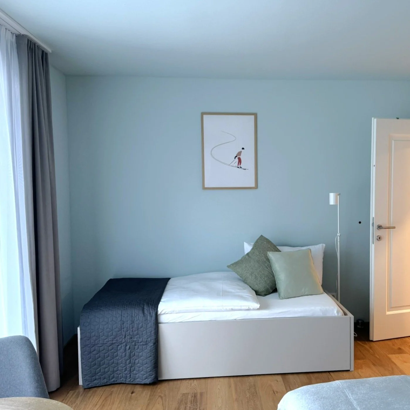 Ein modern eingerichtetes Schlafzimmer mit einem Bett, das mit weißen, grauen und grünen Kissen sowie einer dunklen Decke bedeckt ist. Es gibt weiße Wände, einen Bilderrahmen an der Wand und eine weiße Stehlampe neben der Tür.