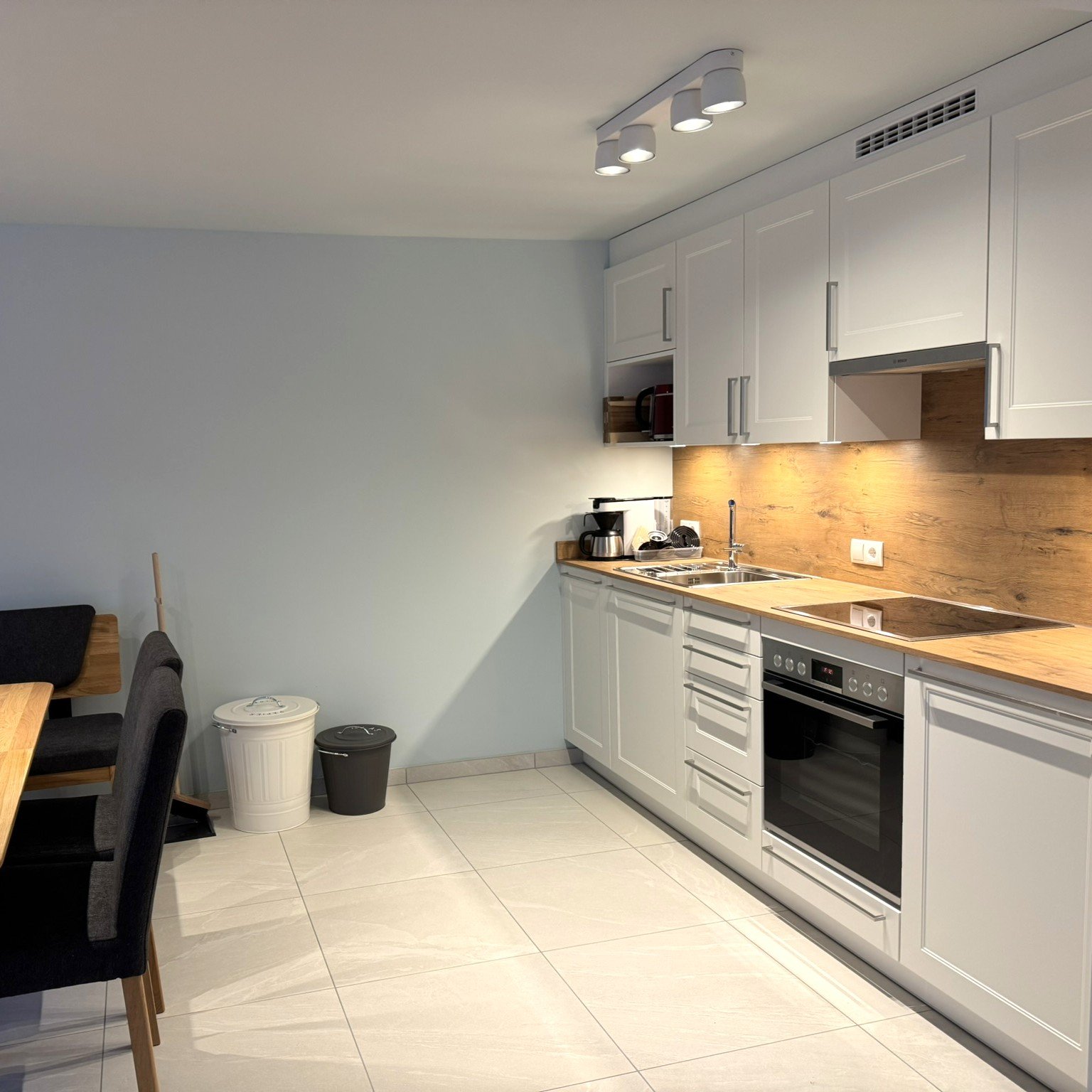 Küche mit weißen Schränken, Holz-Arbeitsplatte, Herd, Backofen, Spüle, Kaffeemaschine, Mikrowelle, und eine beleuchtete Holzrückwand. Teil eines Essbereichs mit dunklen Stühlen und einem Tisch ist sichtbar.