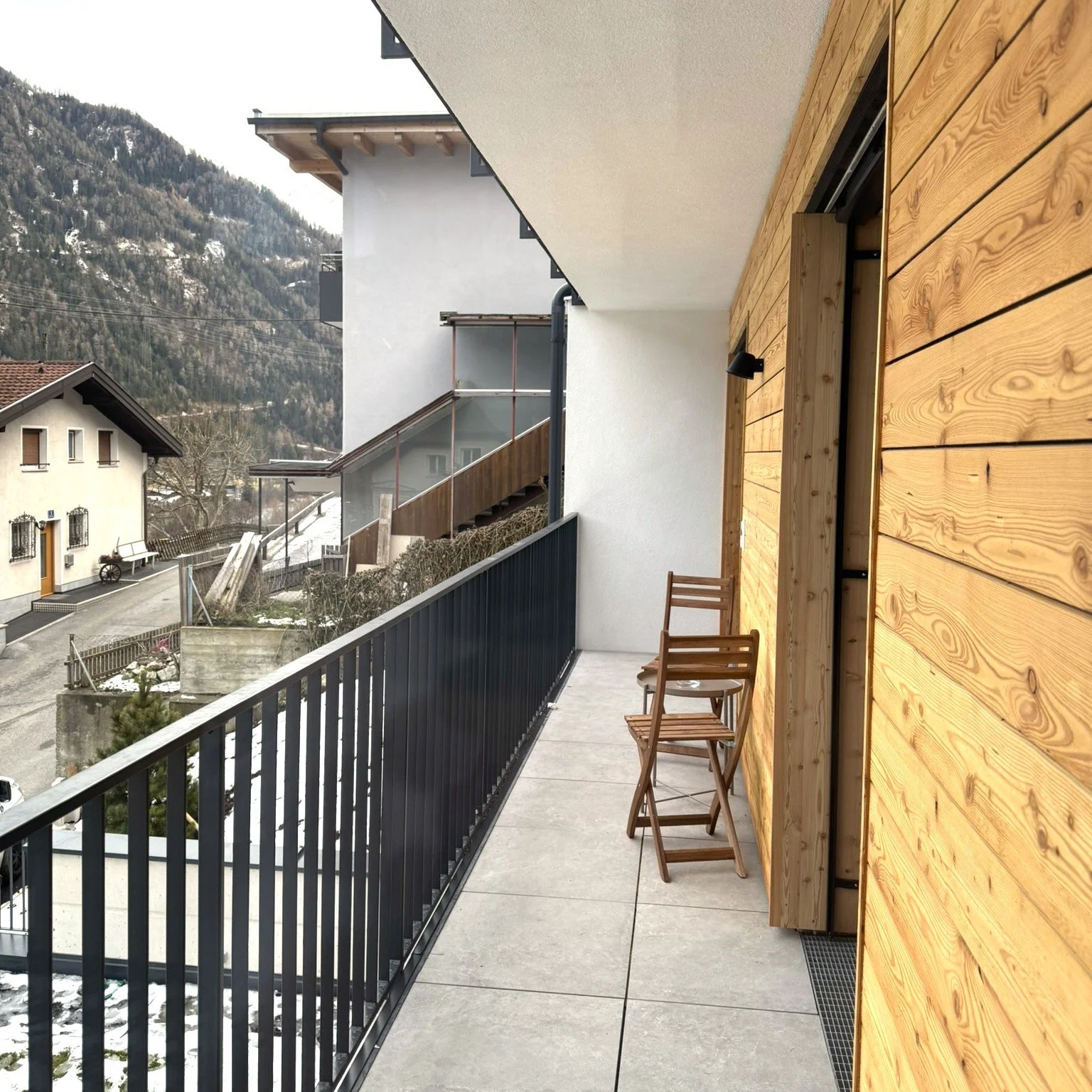 Balkon mit Holzverkleidung, zwei Stühle, schwarze Schutzgeländer, Blick auf Nachbarhäuser und Berge, teilweise Schnee am Boden.