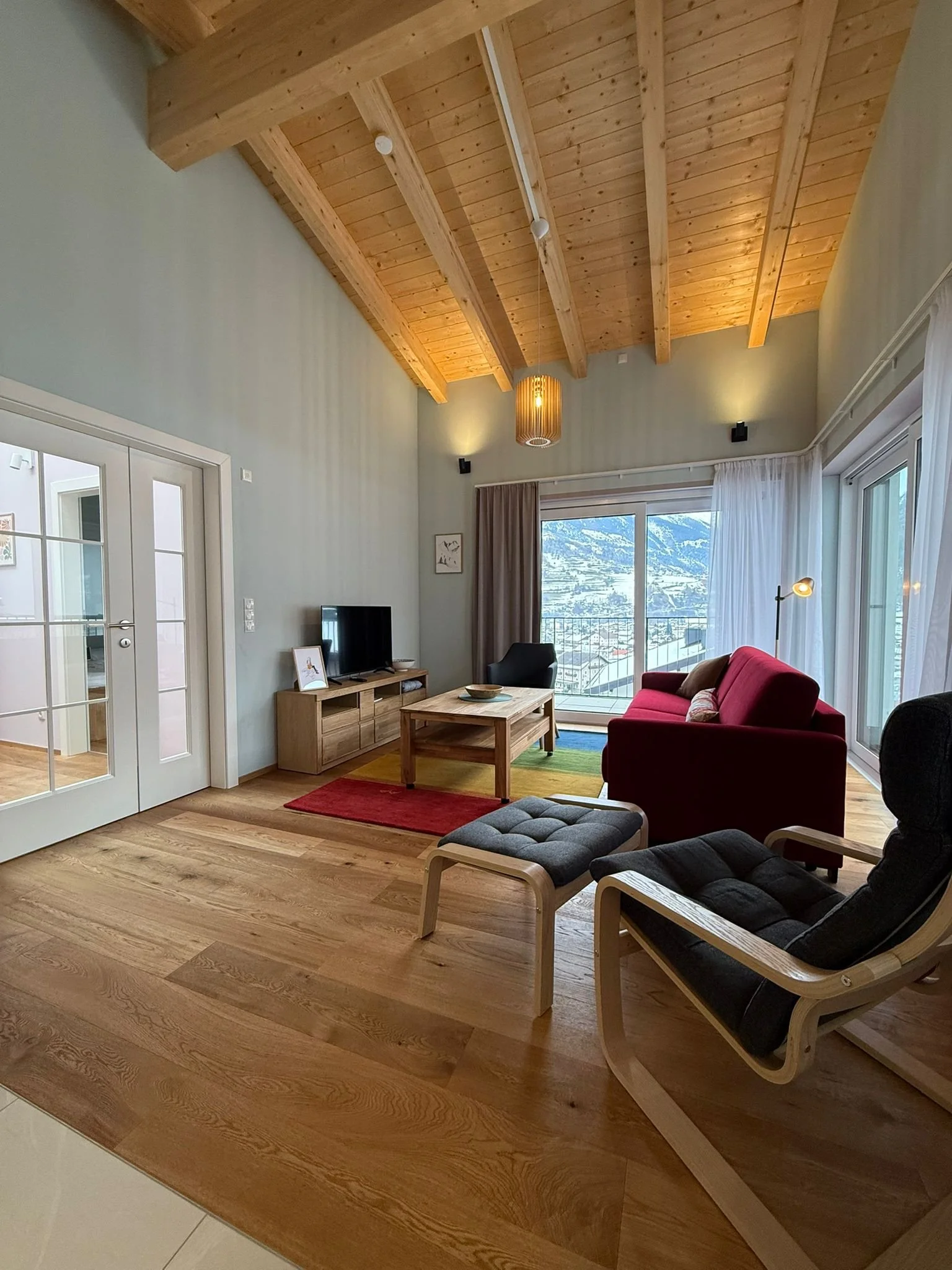 Ein modernes Wohnzimmer mit Holzfußboden, hoher Decke, großen Fenstern mit Tal- und Alpenpanorama, rotem Sofa, schwarzen Sessel, einem kleinen Tisch, einem TV und bunten Teppichen.