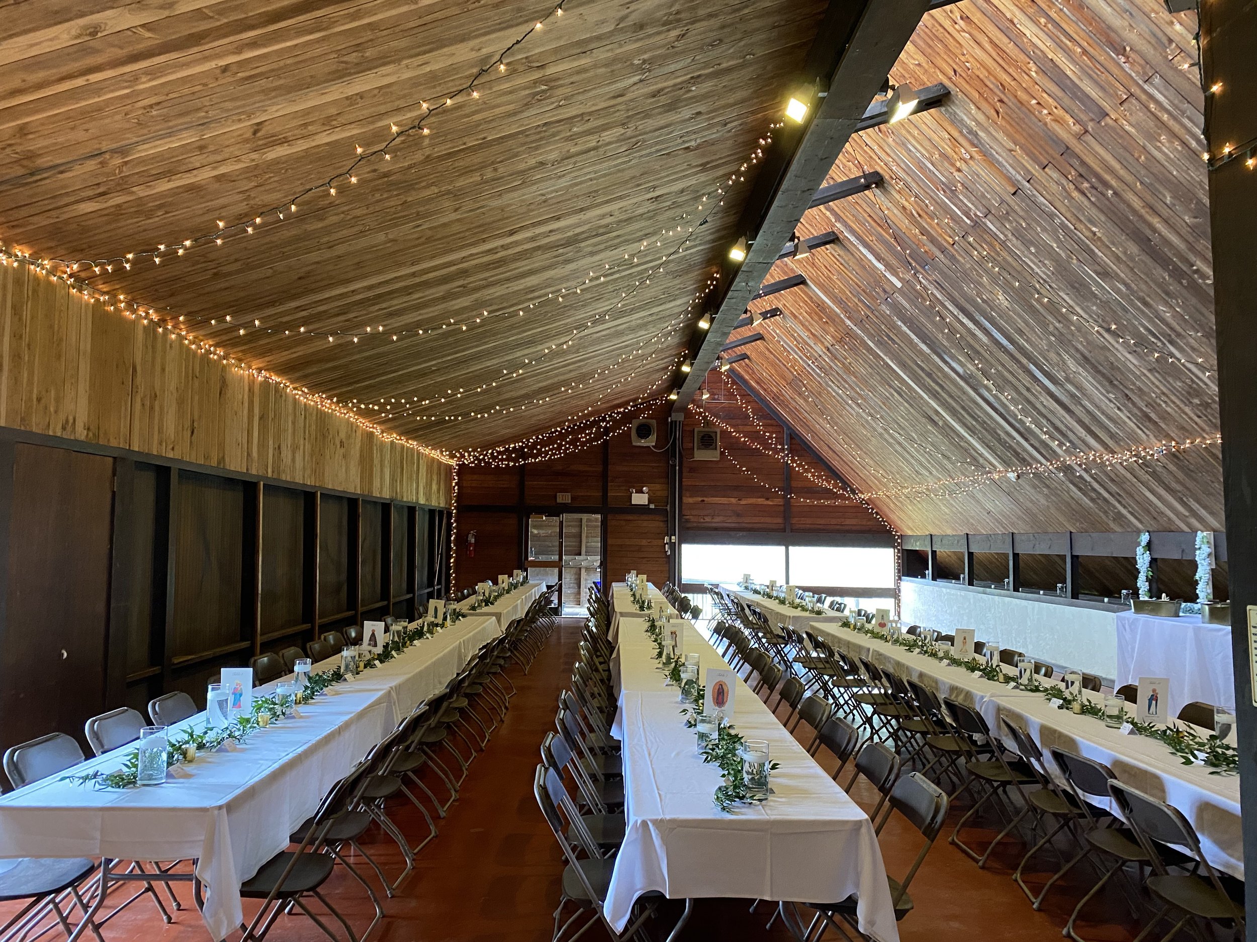 Long banquet tables set with white tablecloths, glassware, and greenery centerpieces inside a wooden hall decorated with string lights
