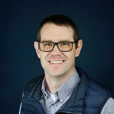 A man with short dark hair and glasses smiling, wearing a light gray shirt and a dark blue vest, against a dark background.