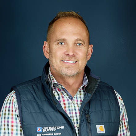 Headshot of a middle-aged man with short reddish hair and blue eyes, smiling, wearing a colorful checkered shirt and a navy vest with a logo, against a dark blue background.
