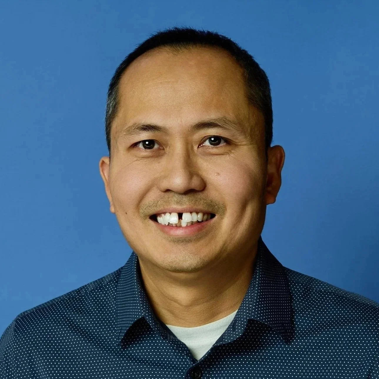 Portrait of a man with short black hair and brown eyes, smiling, wearing a navy blue, dotted shirt against a blue background.