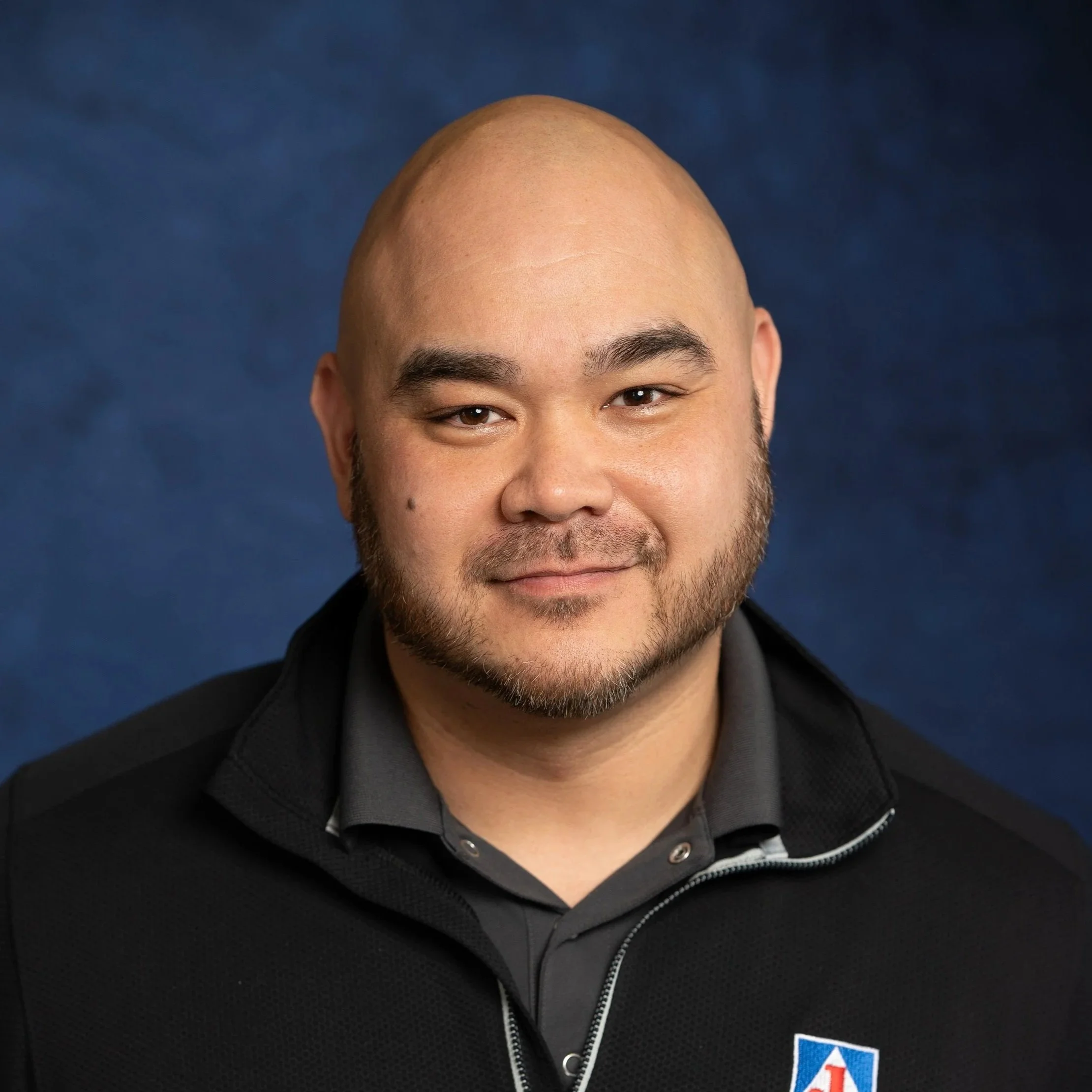 Headshot of a smiling Asian man with a shaved head and beard, wearing a black jacket with a logo on it, against a blue background.