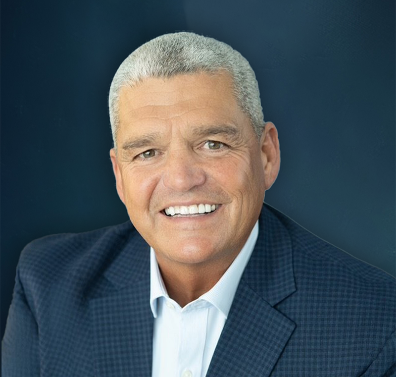 Smiling middle-aged man with short gray hair, wearing a dark blue checkered suit jacket and white shirt, against a dark blue background.