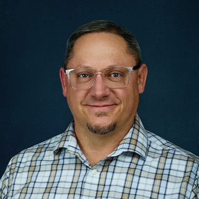 Portrait of a man wearing glasses and a plaid shirt, smiling against a dark blue background.