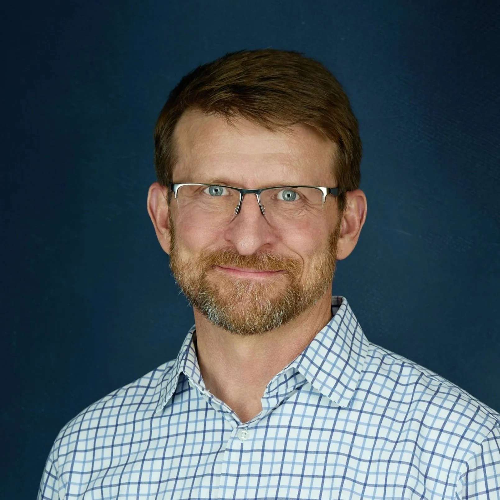 Headshot of a middle-aged man with brown hair, glasses, a trimmed beard, wearing a checkered shirt, against a dark blue background.