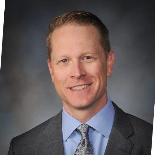 Professional headshot of a smiling man in a suit and tie, against a dark background.
