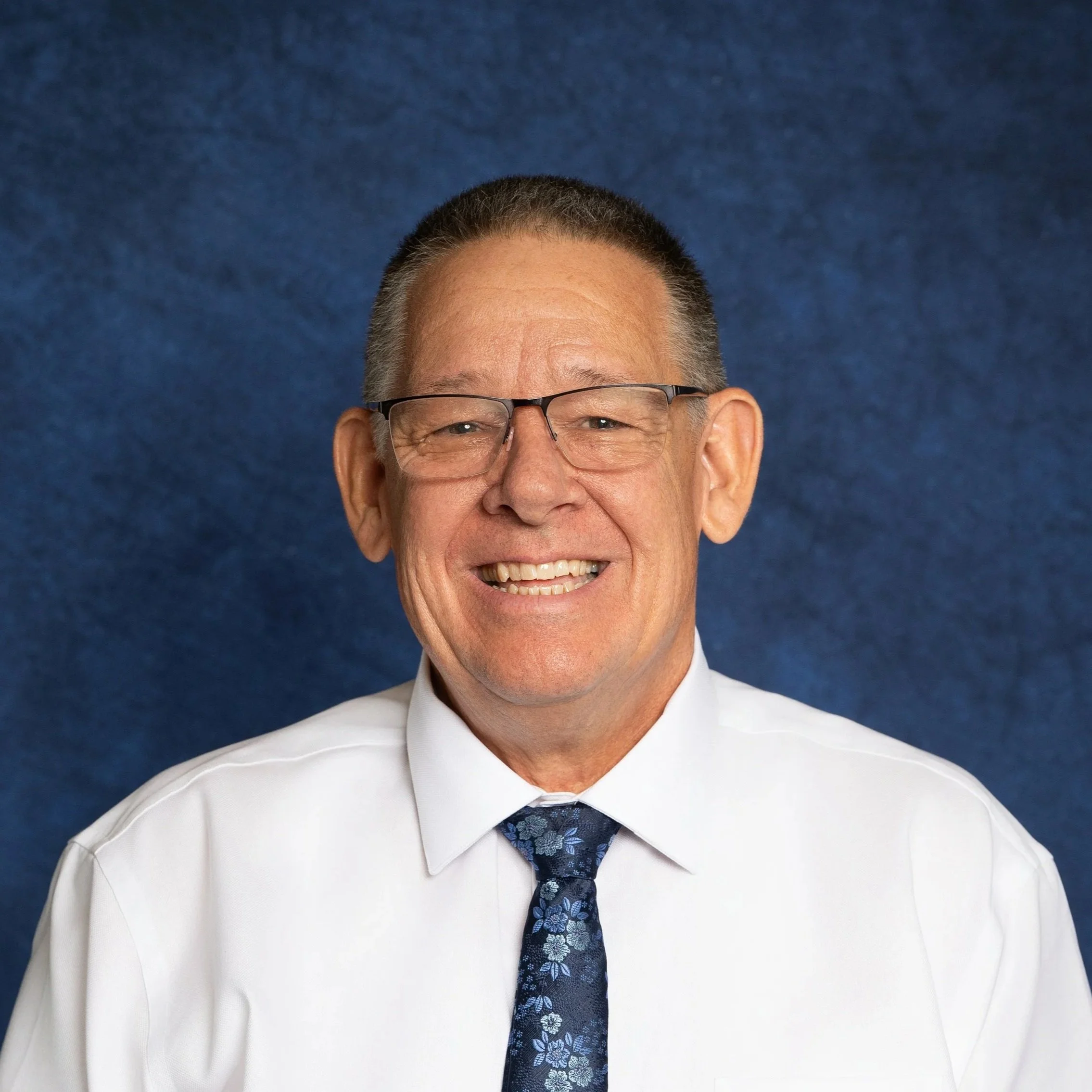 A smiling middle-aged man wearing glasses, a white shirt, and a blue floral tie against a blue background.