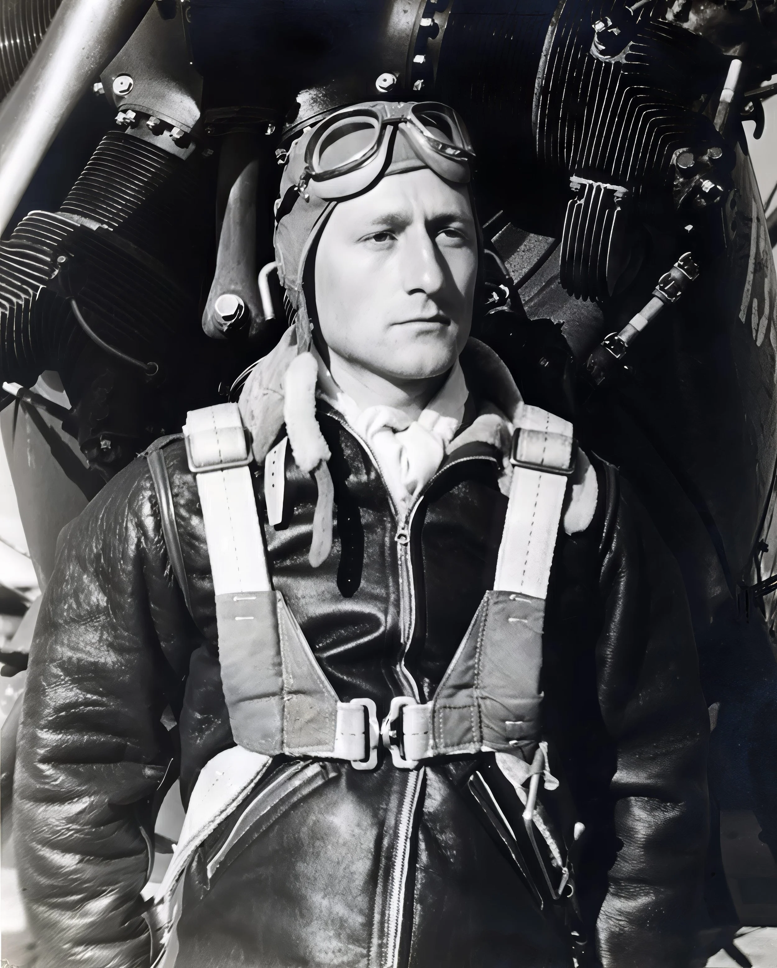 A black and white photo of a man wearing a leather flight jacket, a harness, and goggles, standing in front of an aircraft engine.