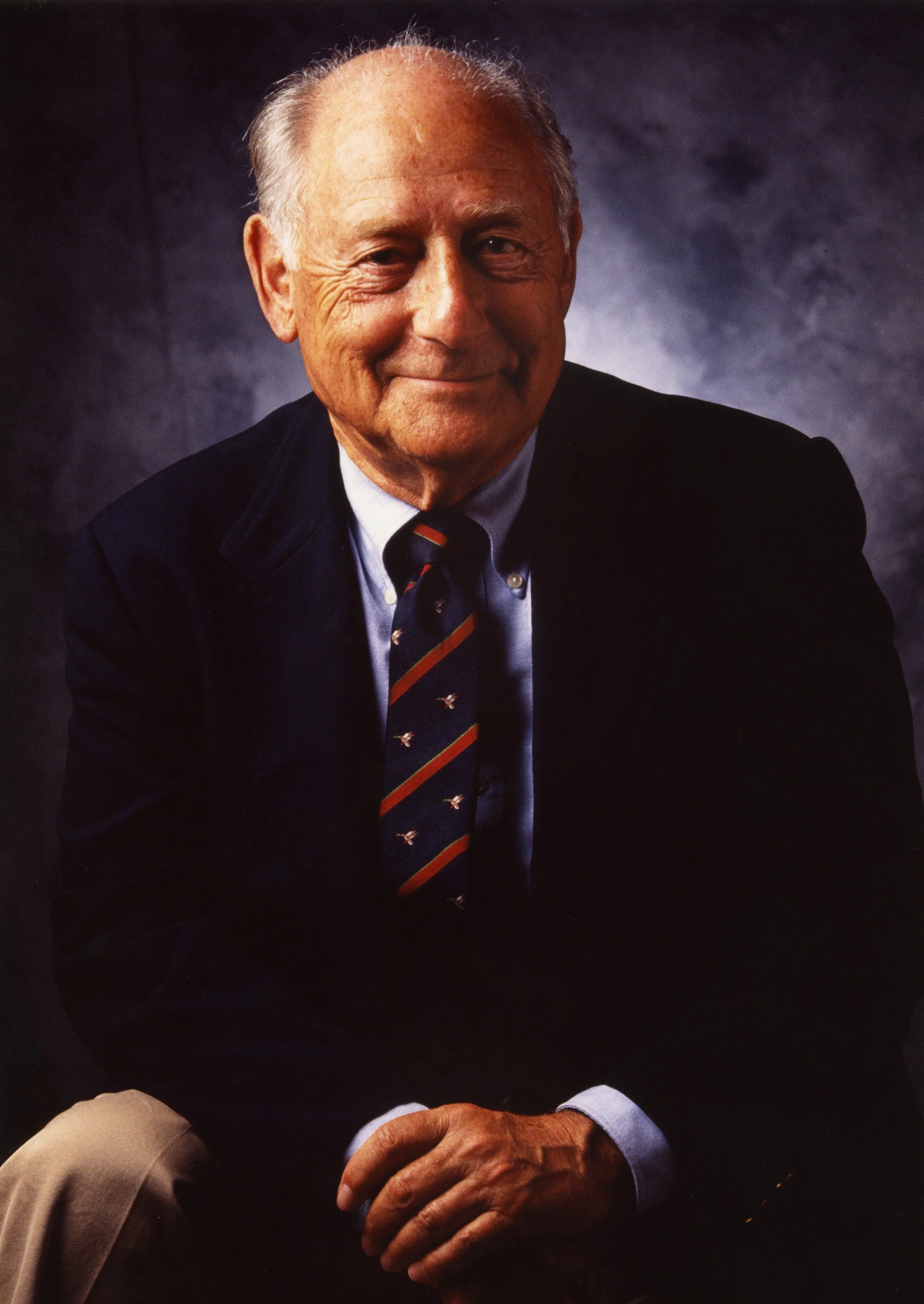 Portrait of an elderly man in a suit, seated with hands clasped, smiling, against a dark mottled background.