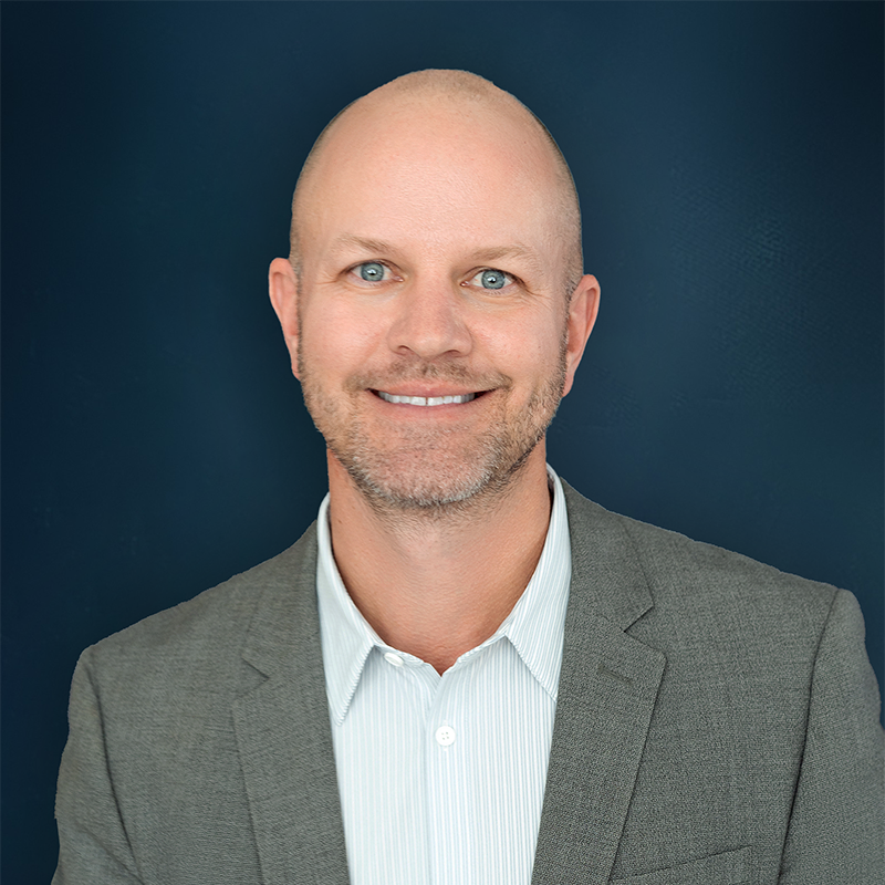 A professional headshot of a smiling bald man with blue eyes, wearing a gray suit jacket and a white collared shirt, set against a dark blue background.