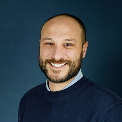 A man with a bald head and beard smiling, wearing a navy blue shirt, against a dark blue background.