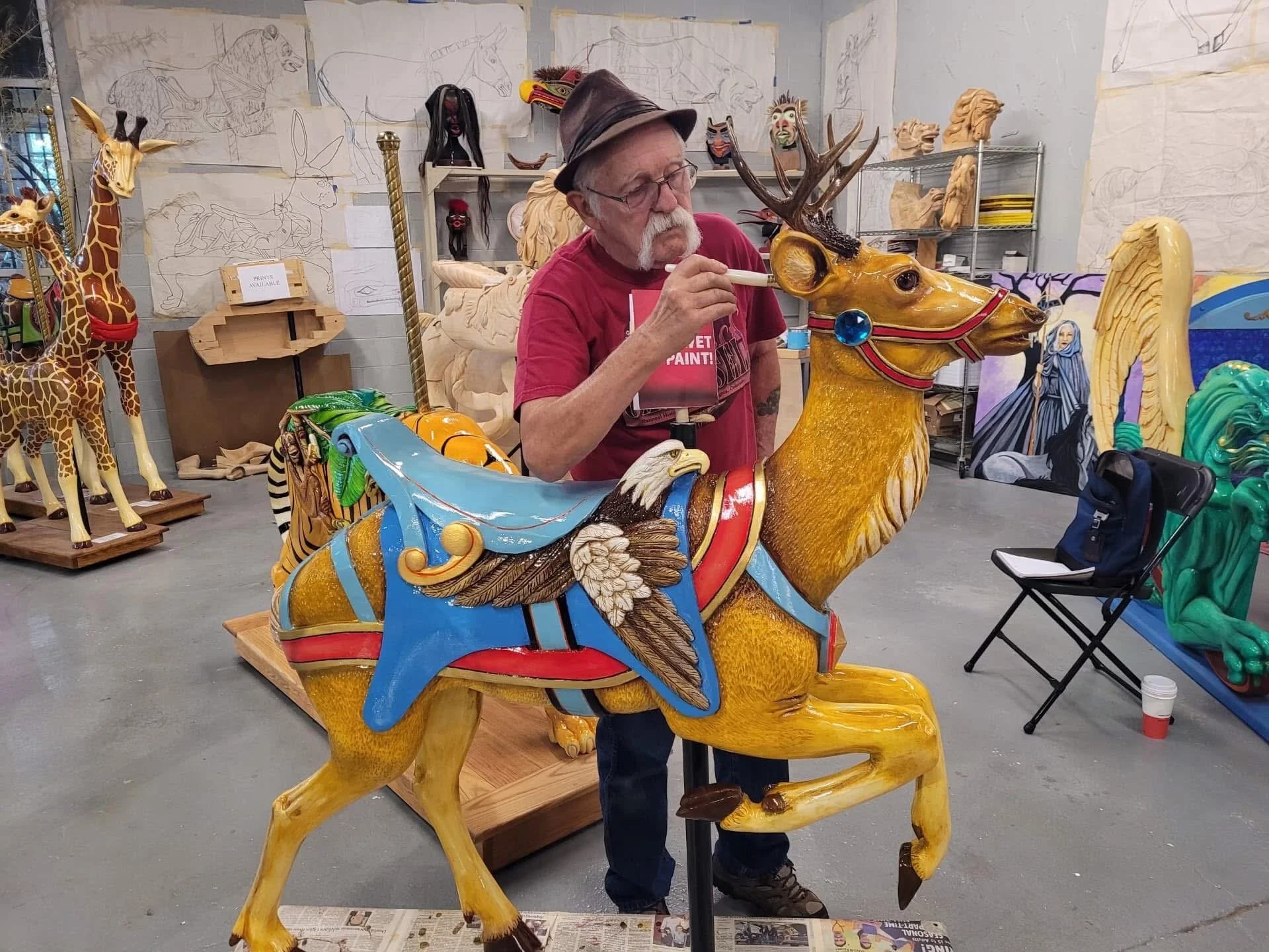 An artist painting a large, colorful carousel animal that resembles a fairy tale creature with the body of a yellow dog, the head of a deer, and wings, in a workshop filled with other animal sculptures and sketches.