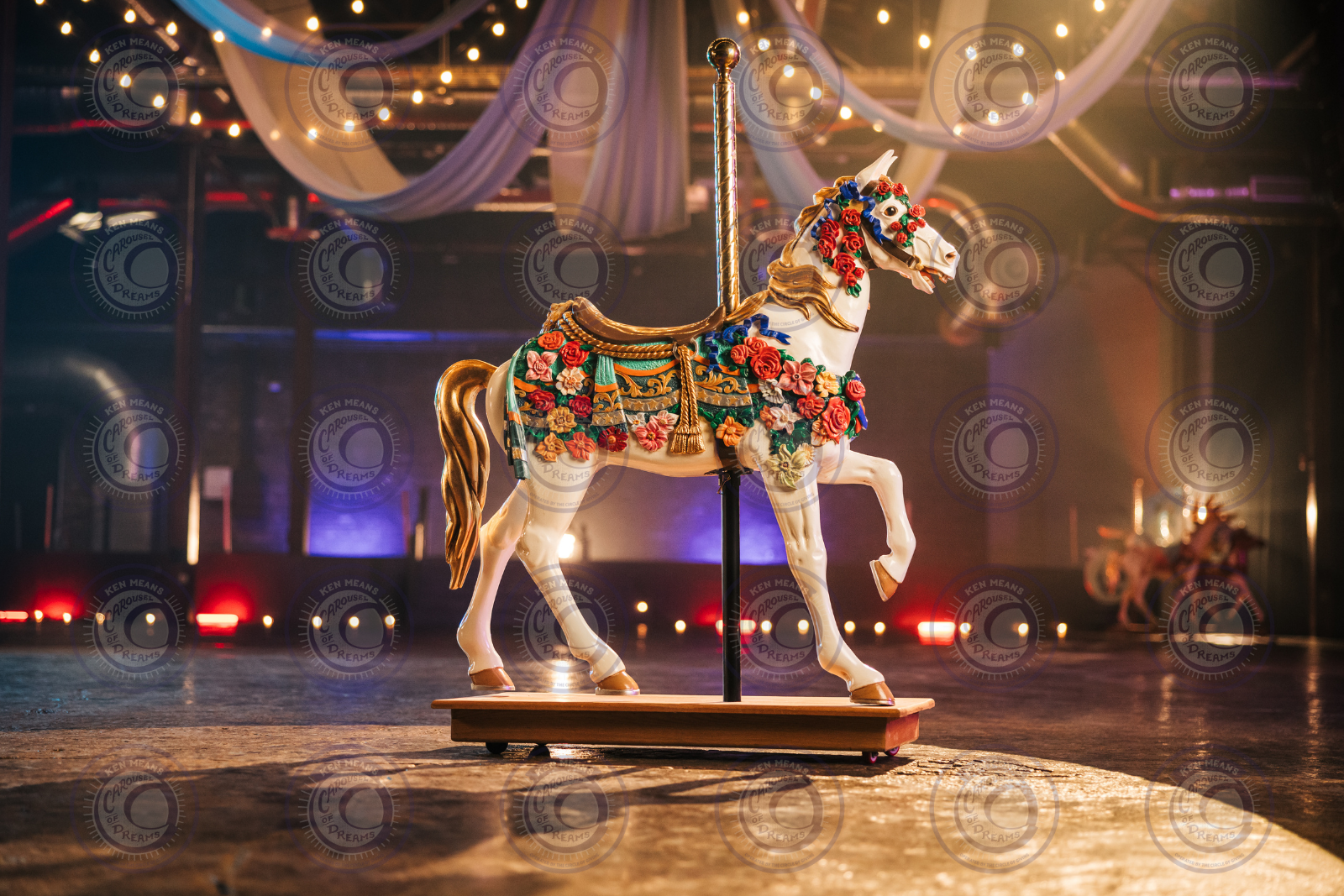 A carousel horse adorned with colorful flowers and decorations, under hanging lights and draped fabric in a dimly-lit setting.