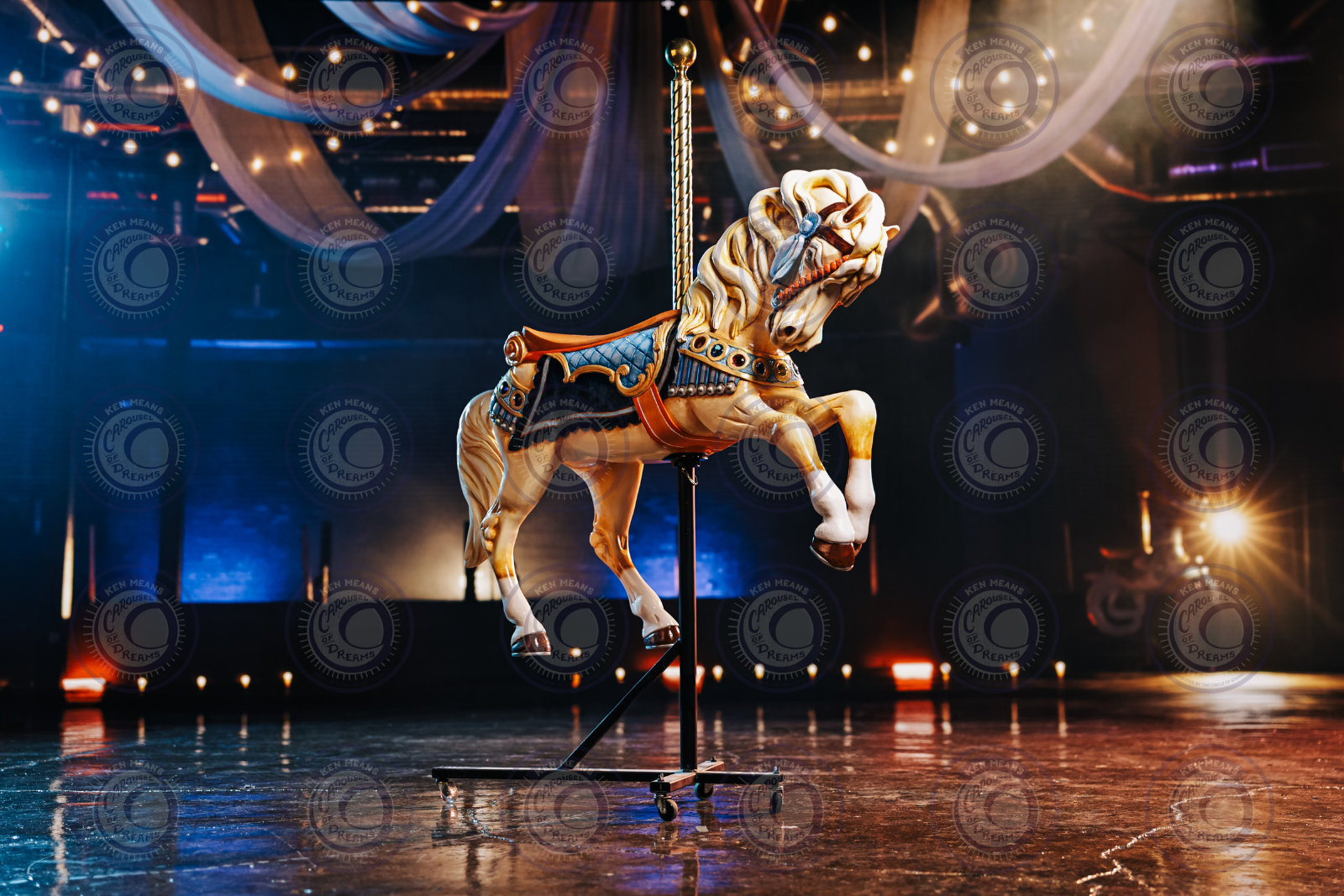 A detailed vintage carousel horse in an indoor setting with decorative lights and draped fabric hanging from the ceiling. The horse is mounted on a stable support stand with wheels.