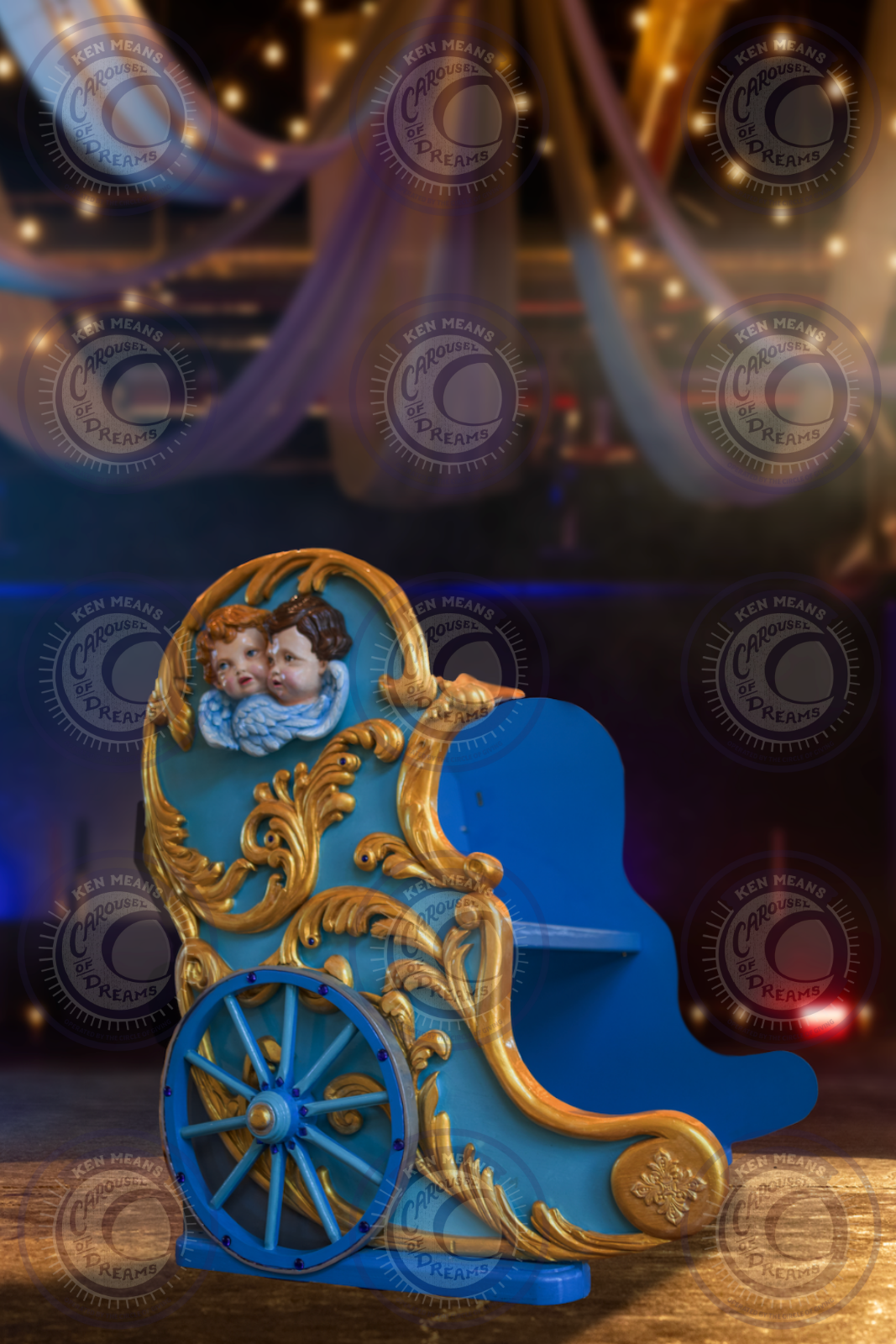 A colorful toy carriage with gold decorative details and two toy cherubs on top, placed on a wooden surface in a dimly lit room with draped fabric and string lights in the background.