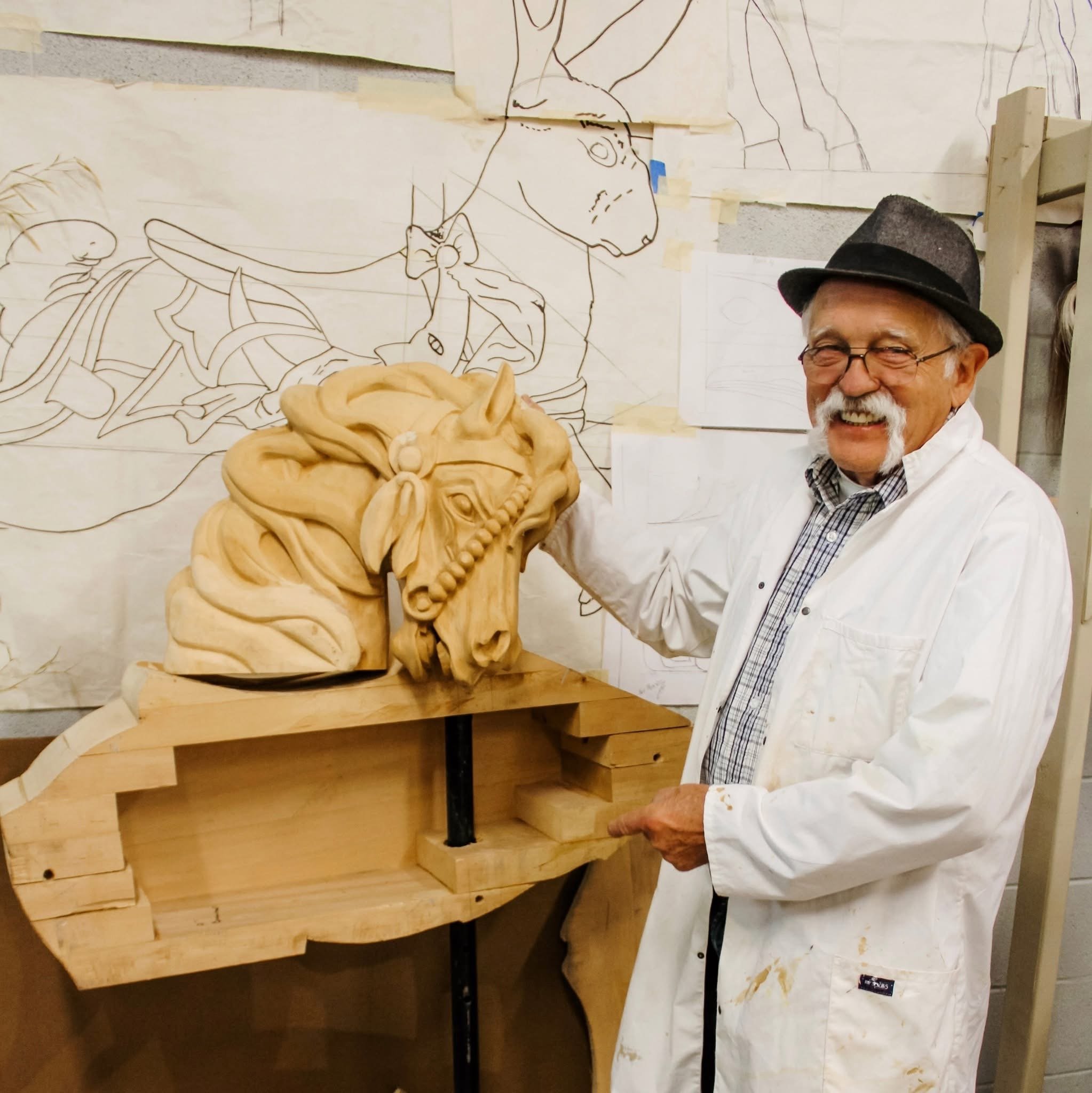 An older man with glasses, a hat, and a white coat is smiling while holding a large wooden sculpture of a lion's head. The background features sketches of animals, including a horse and a deer, taped to the wall.
