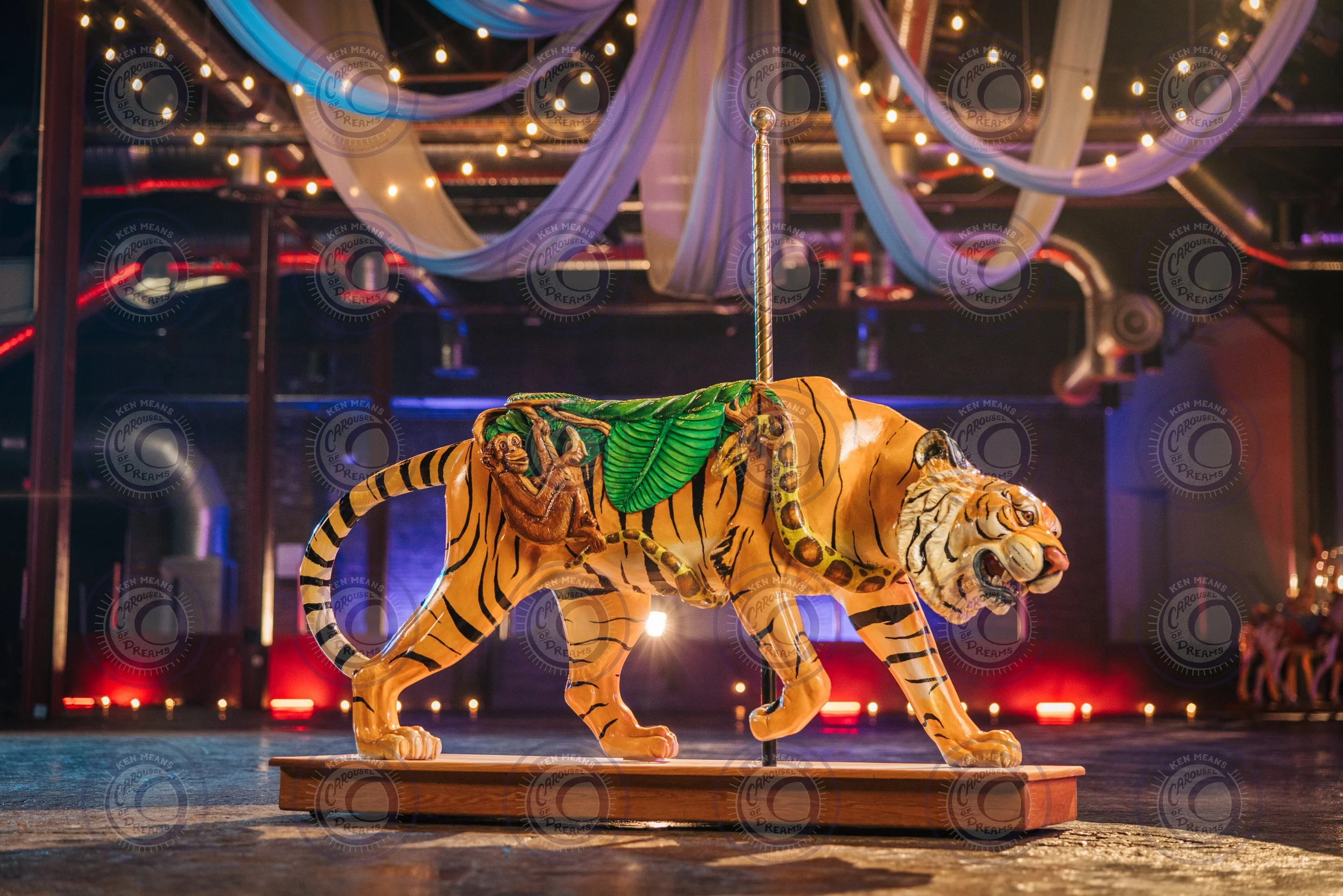 A wooden tiger carousel figure with a monkey and snake design in a brightly lit indoor setting with draped fabric and string lights.