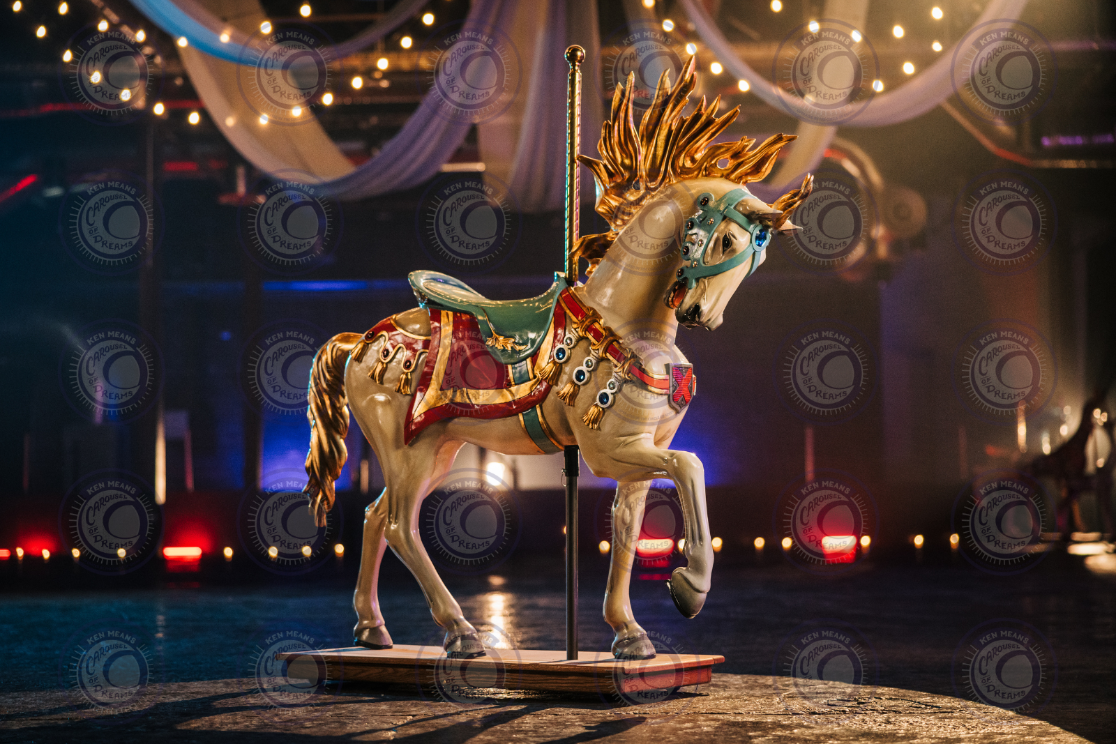 Colorful carousel horse with golden mane and tail, decorated with red and green saddle and harness, mounted on a wooden platform, inside a circus tent with draped fabric and string lights.