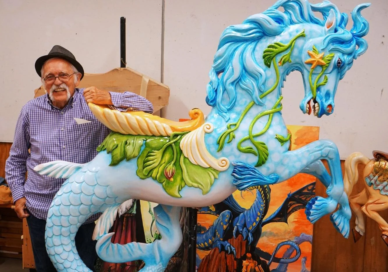 An elderly man with glasses, a gray beard, wearing a straw hat and checkered shirt, smiling and holding onto a large, colorful, fantasy-style dragon sculpture with a blue mane, scales, and leafy decorations.