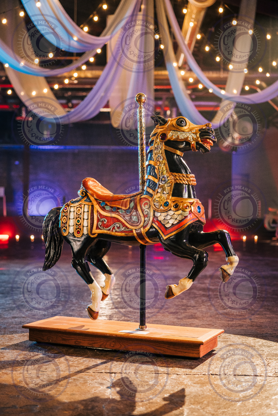 Decorative black carousel horse with colorful ornaments on a wooden base, surrounded by draped fabric and string lights in a dimly lit room.