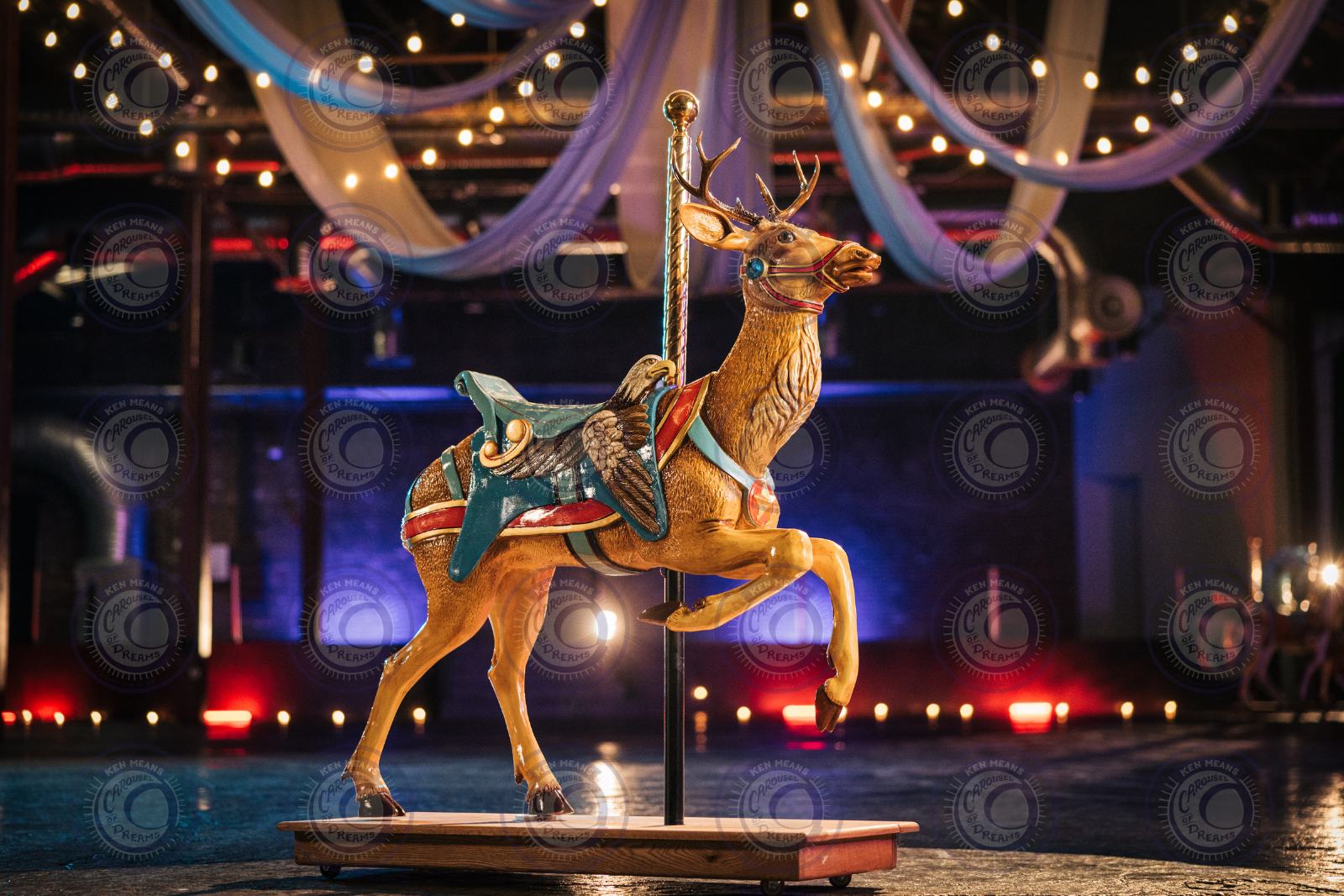 Decorative carousel deer with colorful saddle, set in an indoor venue with string lights and draped fabric above.