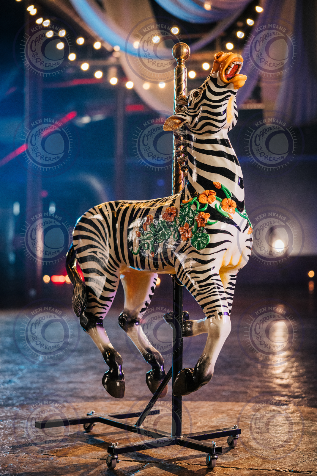 A colorful carousel zebra with a black and white stripe pattern, decorated with orange flowers and green leaves, standing on a metal stand with decorated string lights and a draped fabric background.
