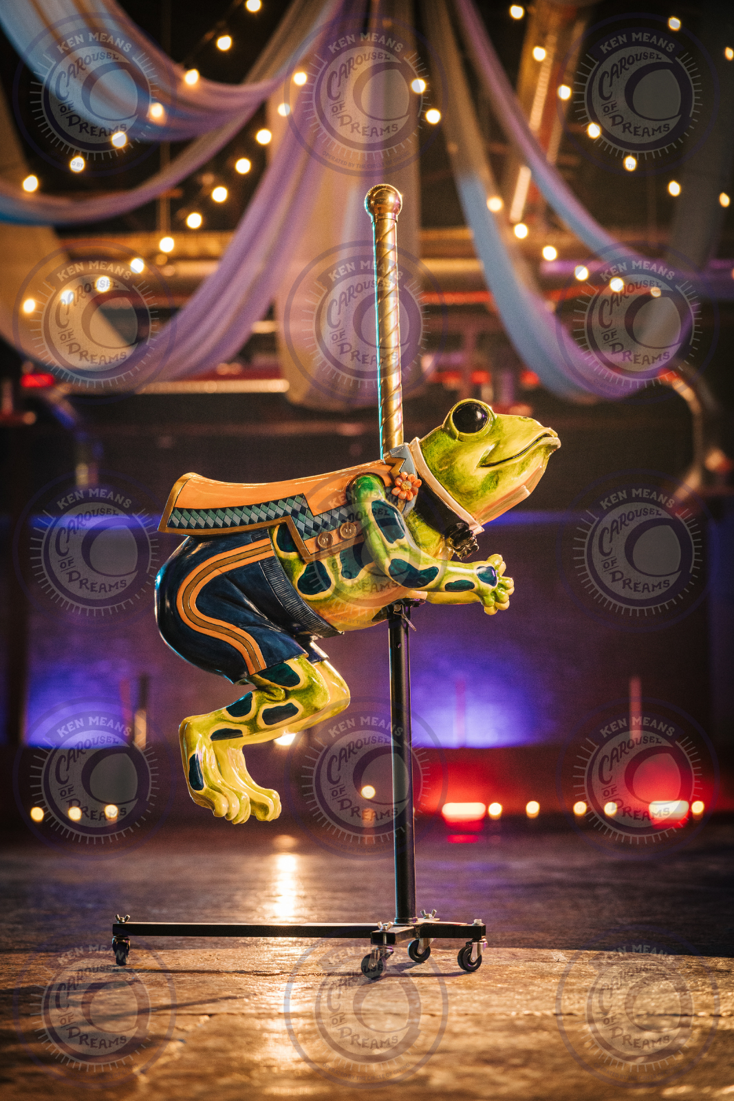A colorful frog sculpture resembling a carousel ride, mounted on a wheeled stand, with draped strings of lights in the background. The setting appears festive and illuminated.
