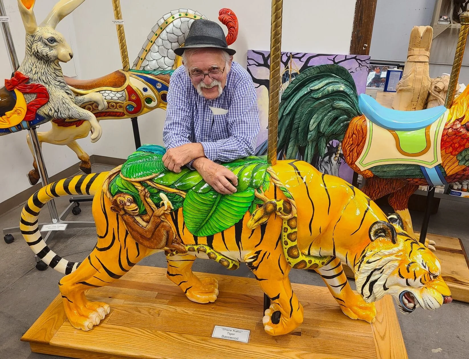 An elderly man with glasses and a hat leaning on a colorful, painted tiger sculpture with jungle animals, including a monkey and a giraffe, on its back. The scene is indoors, with other large painted animal sculptures in the background.