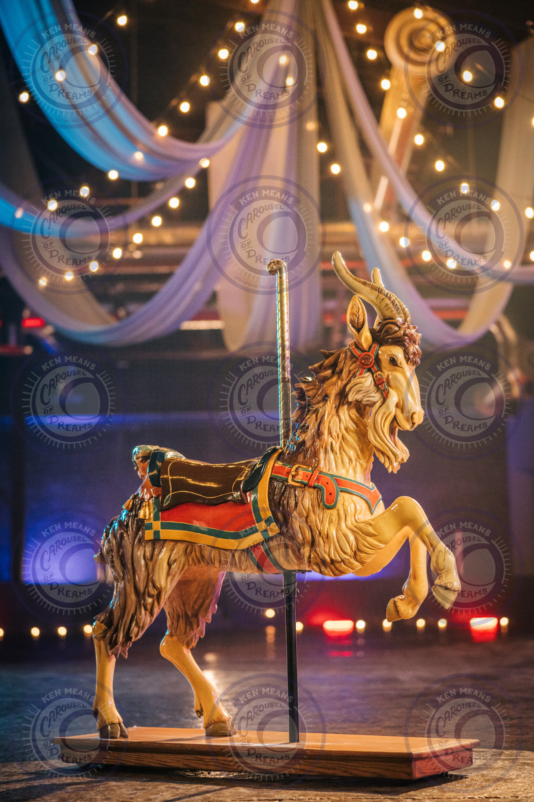 A carousel goat sculpture with ornate design, placed on a stand, set against a background of draped fabric and string lights.