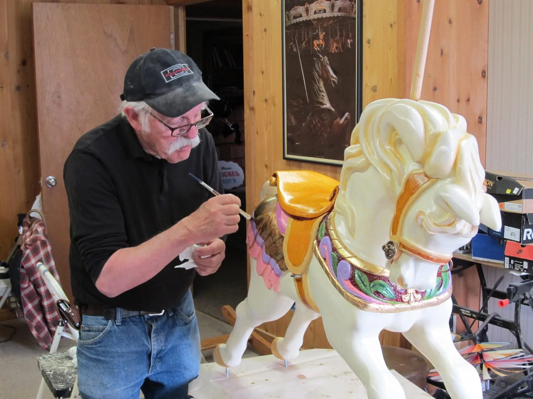 An elderly man with glasses and a cap painting a carousel horse sculpture in a workshop.