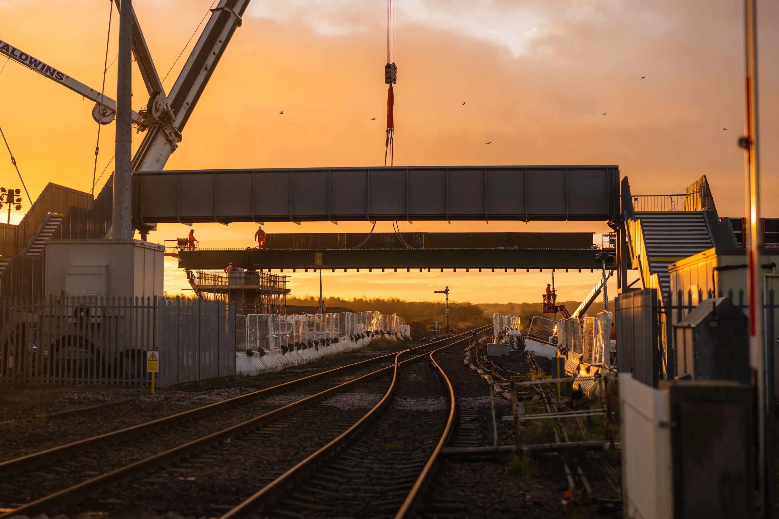 Train tracks with newly installed bridges as the sun rises, giving an orange, glowing sky. Birds can be seen flying in the sky as construction workers carry out work above and below