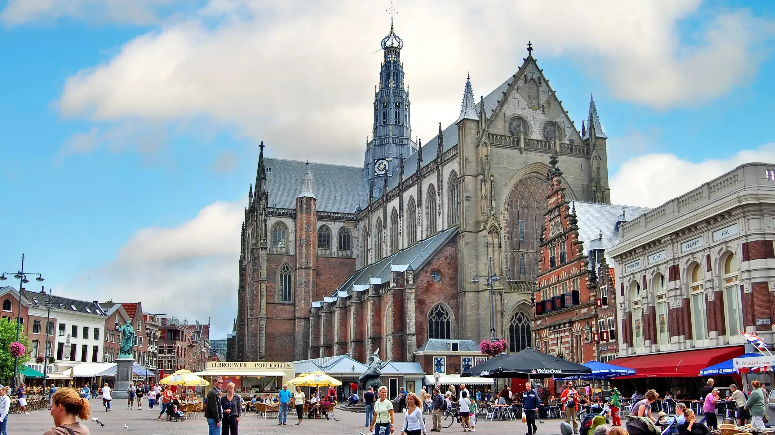 Een druk stadsplein met mensen, terrasjes met parasols, een stand met een gouden banner, en een grote gotische kerk op de achtergrond, onder een deels bewolkte hemel.