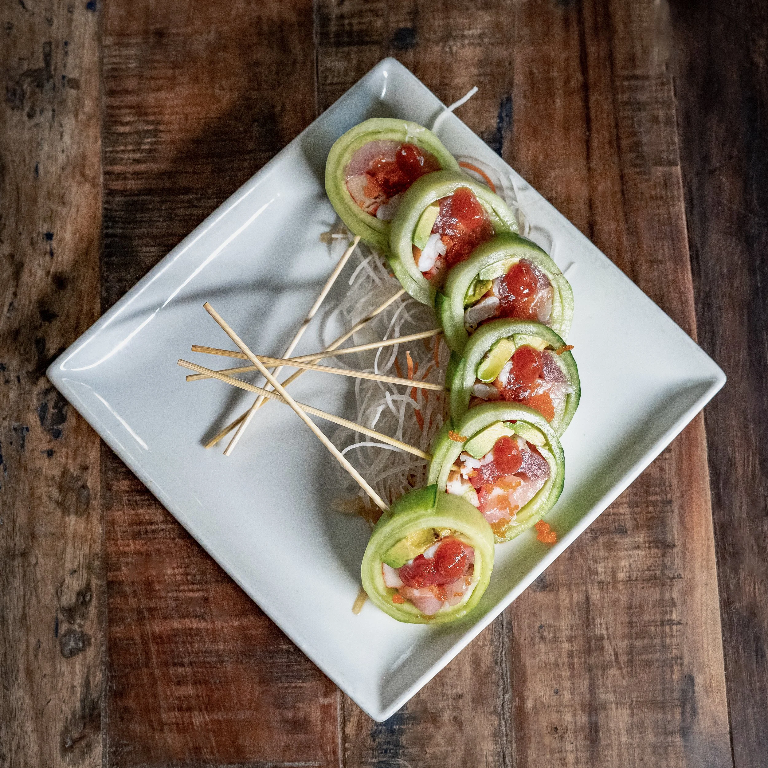 Sliced cucumber rolls filled with seafood and topped with sauce, served on a white rectangular plate with skewers and shredded vegetables on a wooden surface.