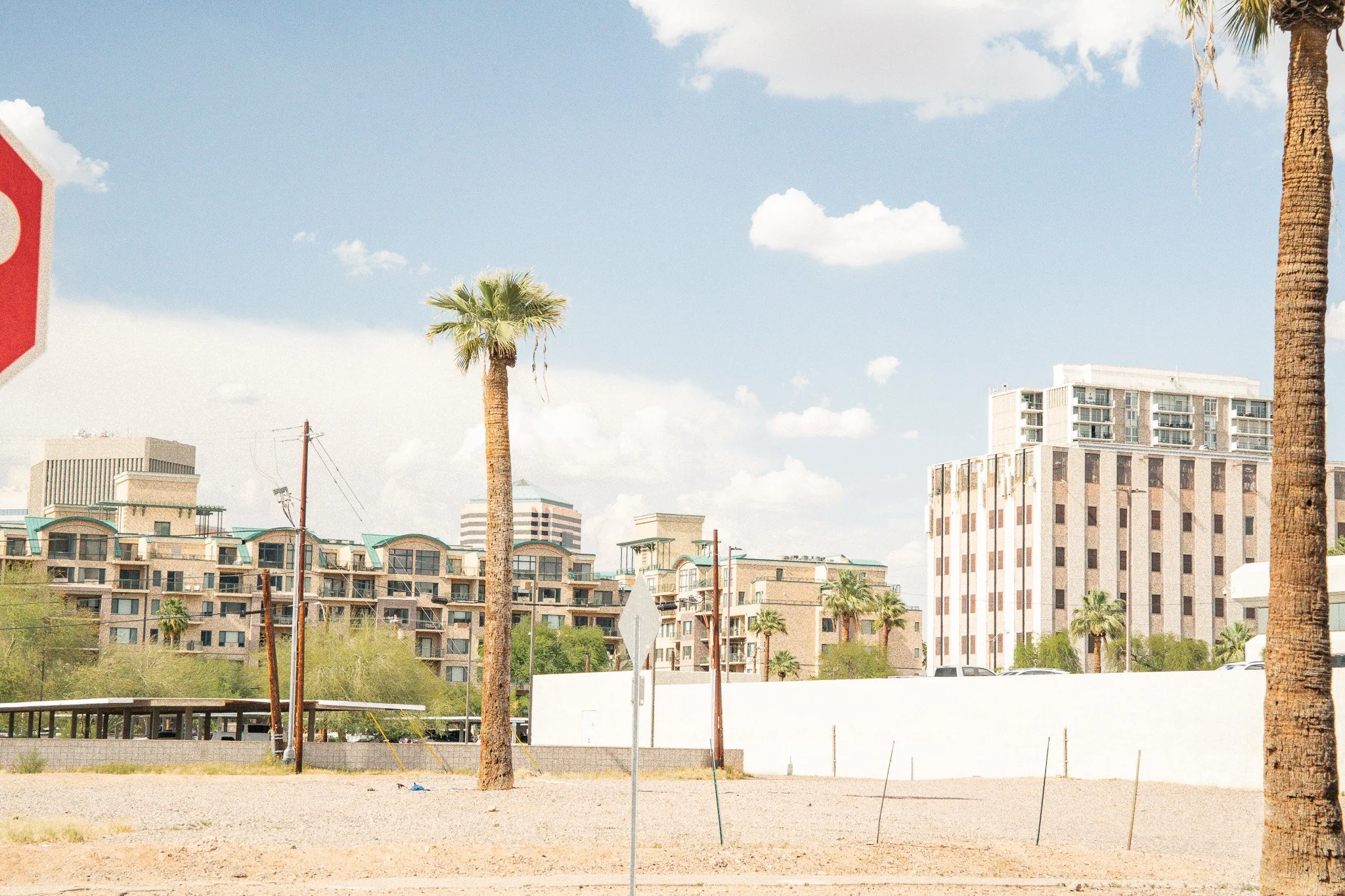 Cityscape with palm trees, multi-story buildings, a white wall, parked cars, utility poles, and a stop sign in a sunny, partly cloudy sky.