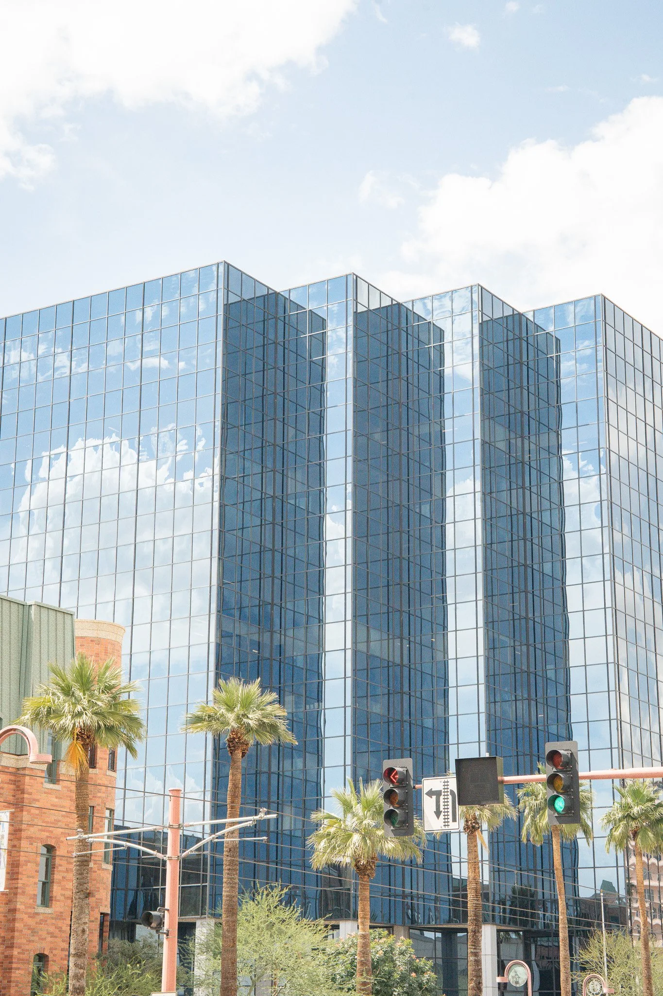 Modern glass office building reflecting the sky and clouds, with palm trees and traffic lights in the foreground.