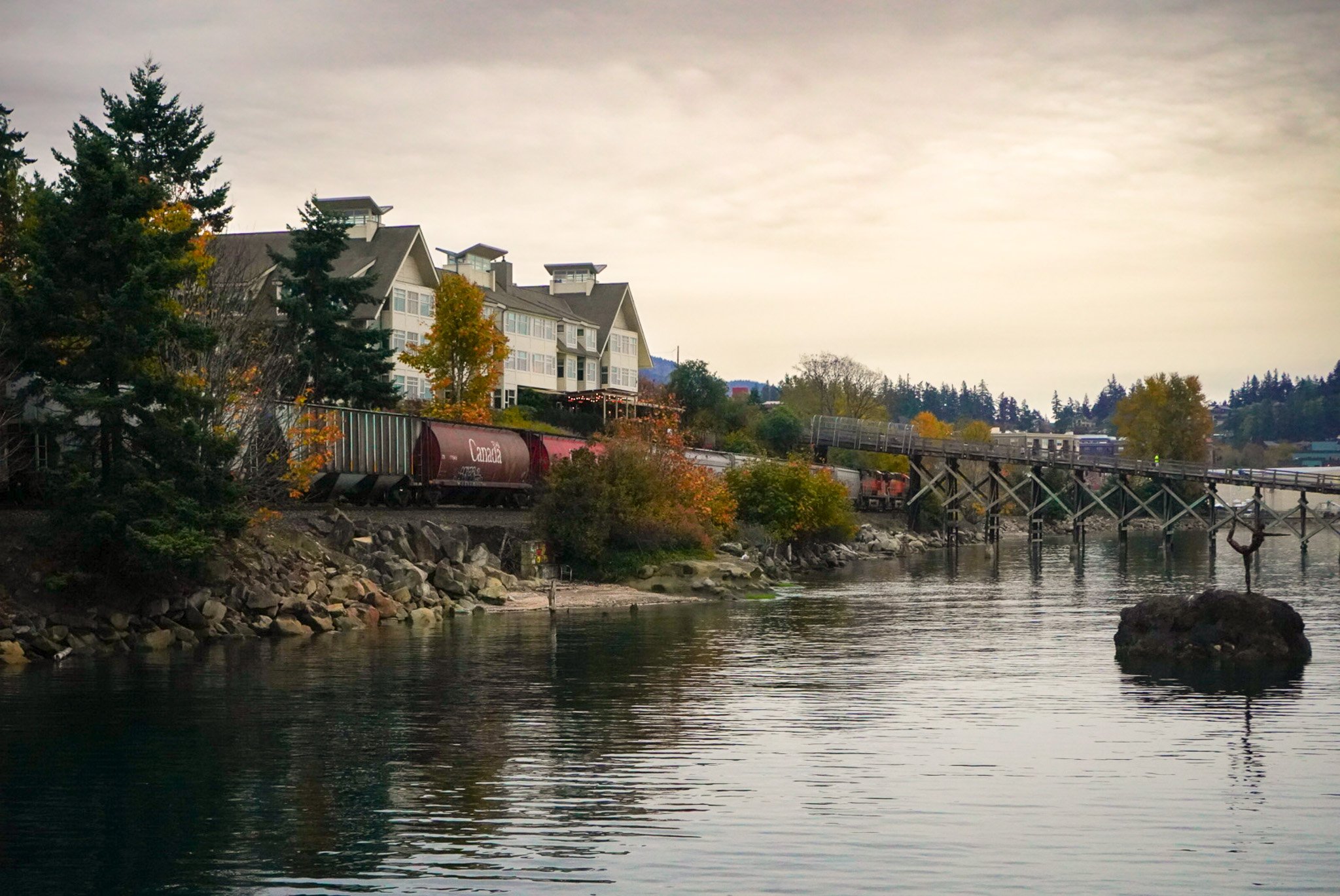 A riverside scene with a train passing over a bridge, houses on a hillside, trees, and calm water reflecting the sky.