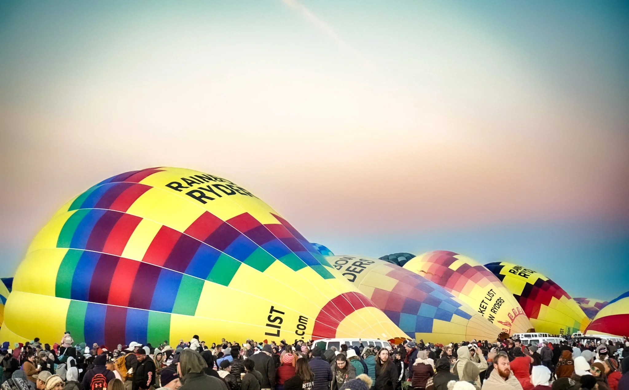 A large crowd watching hot air balloons being inflated, including multiple colorful balloons with yellow, red, green, and blue patterns, at a hot air balloon festival during dawn or dusk.