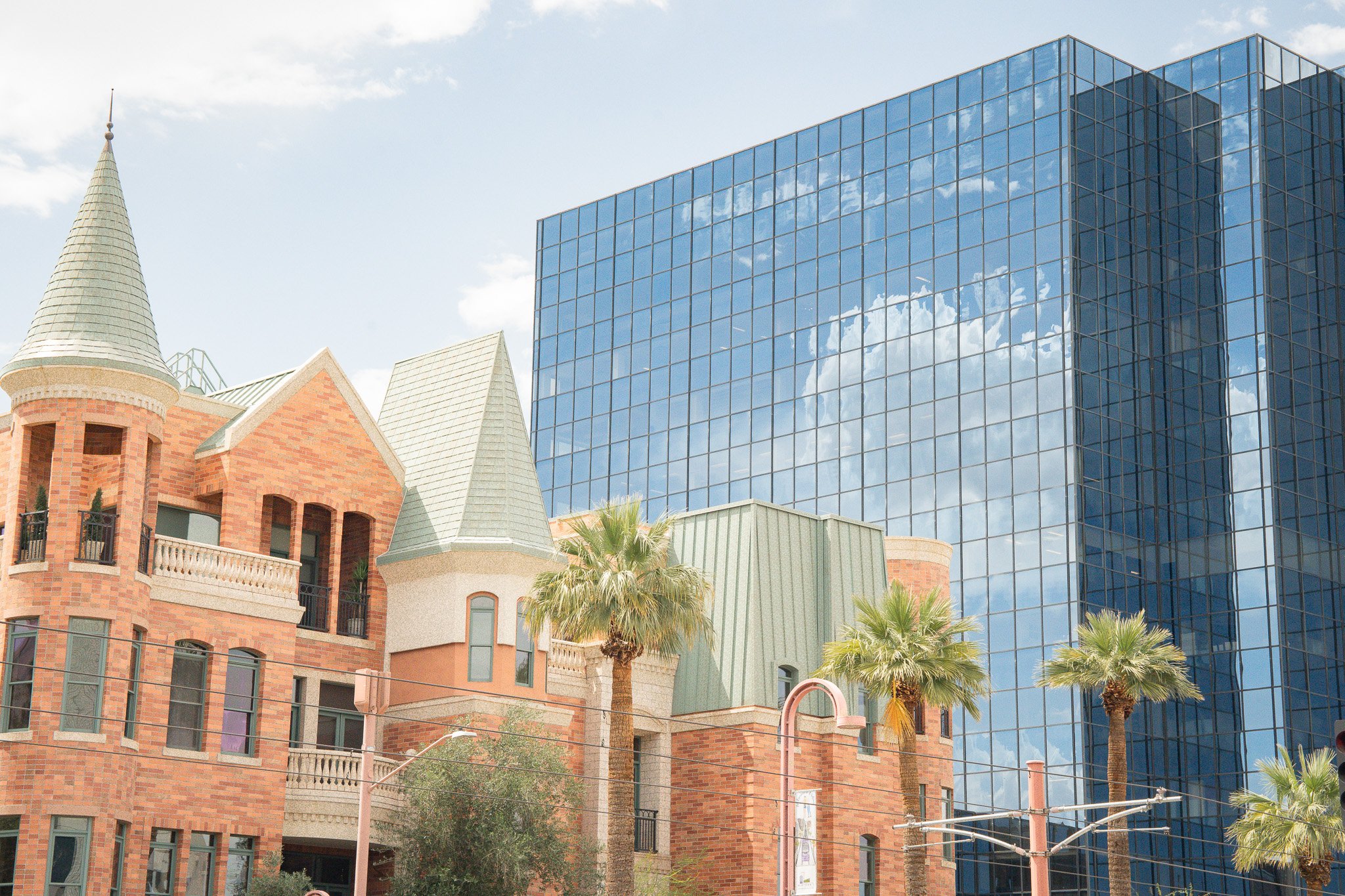 Modern glass building reflecting clouds and sky, adjacent to a building with castle-like architecture and palm trees in an urban setting.