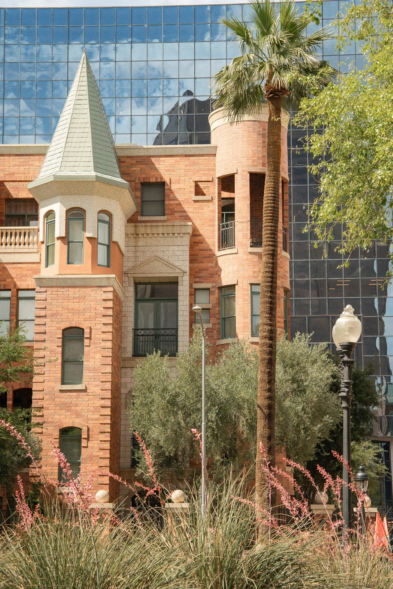 A building with a castle-like design featuring a turret and brick facade, reflected in a modern glass building behind it. There are trees, a street lamp, and some pink flowering plants in the foreground.
