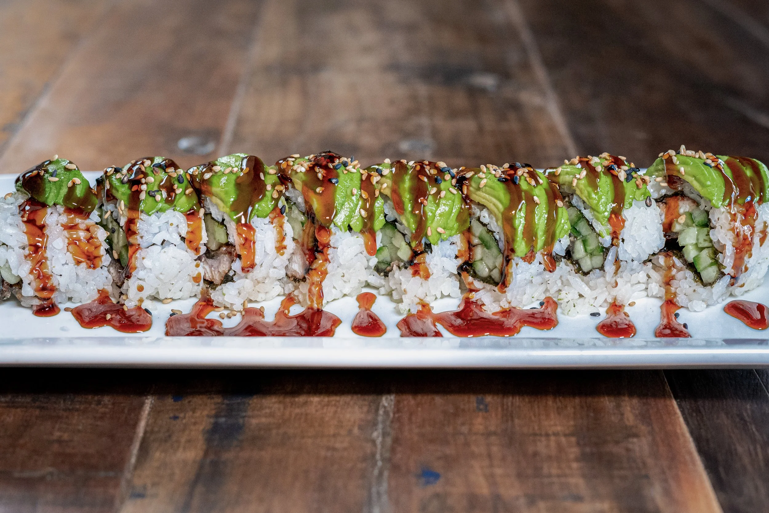 A row of sushi rolls topped with avocado, eel sauce, and sesame seeds on a long white plate with a dark wooden table background.